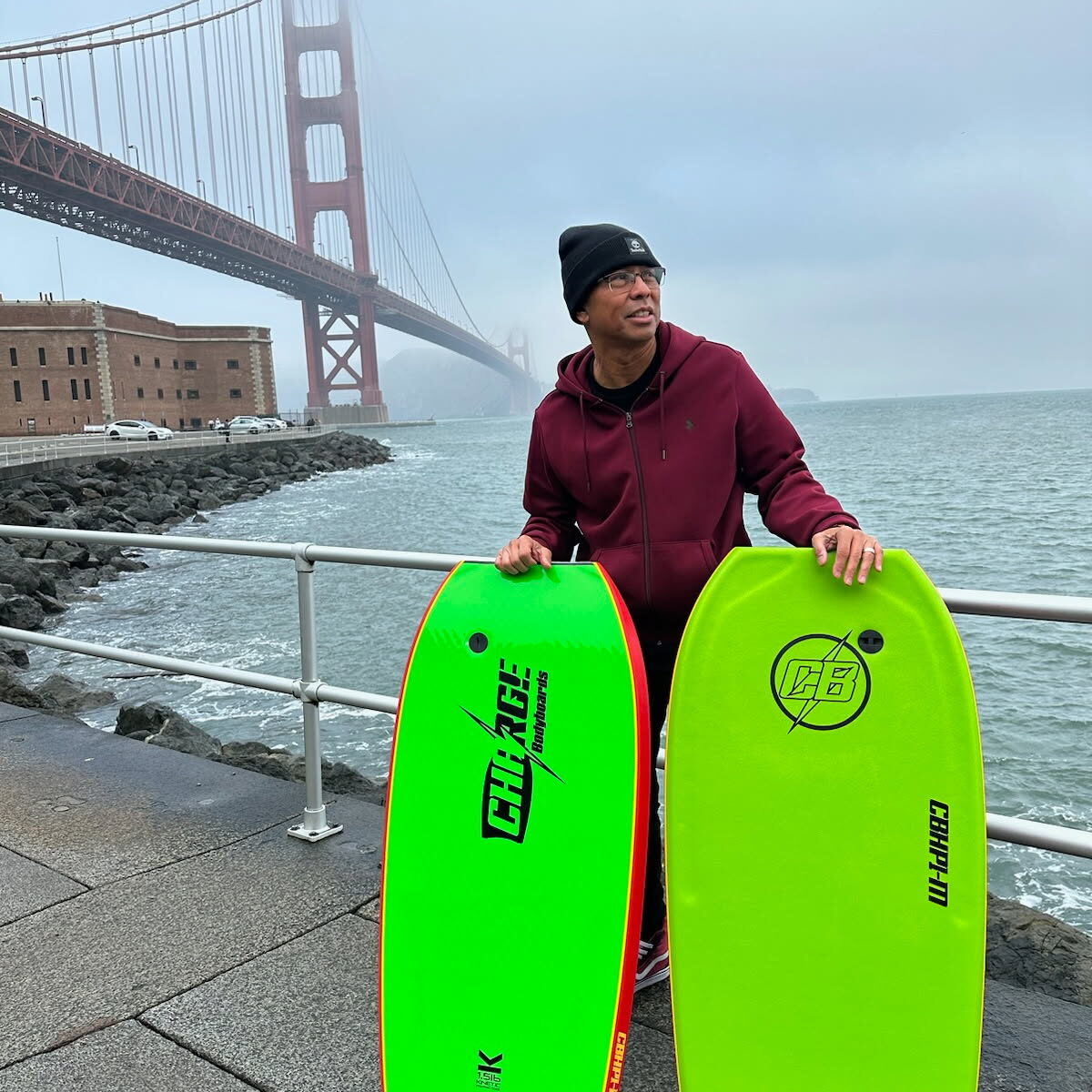 Person holding two bright green bodyboards near the Golden Gate Bridge on a cloudy day.